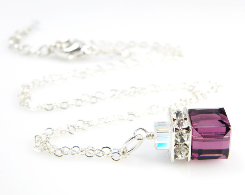May include: A silver chain necklace with a pendant featuring a large, square, purple crystal surrounded by smaller clear crystals.