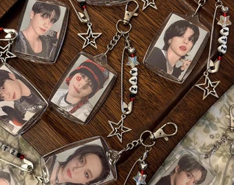 May include: A set of eight clear plastic keychains featuring photos of a K-pop boy band. Each keychain has a silver star charm and a silver safety pin with a beaded chain that spells out "ATZ".