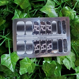 May include: A set of nail wraps in a clear plastic package. The wraps feature a variety of designs, including ombre, geometric patterns, and solid black with leaf accents. The text "Bioactive Nails" is printed on the package.