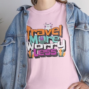 Puede incluir: Camiseta rosa con un estampado gráfico de un gato y el texto "Travel More Worry Less" en letras de colores de estilo 3D.