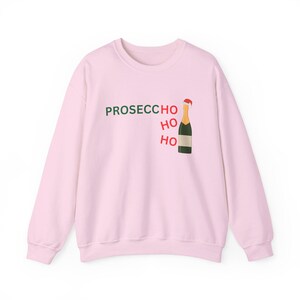 Op de afbeelding: Een lichtroze sweatshirt met de tekst "PROSECCO HO HO HO" in groen en rood. Een cartoon champagnefles met een kerstmuts staat naast de tekst. De sweatshirt heeft een ronde hals en lange mouwen.