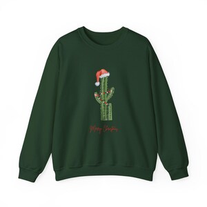 May include: A dark green sweatshirt featuring a cactus decorated with Christmas lights and a Santa hat. The words "Merry Christmas" are written in red script below the cactus design. The sweatshirt is made of a soft material.