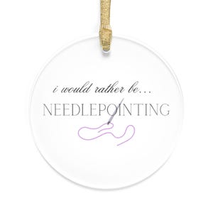 May include: A clear, round ornament with the text "i would rather be... NEEDLEPOINTING" in black and gray. A purple needle and thread design is below the text. The ornament hangs from a gold ribbon.