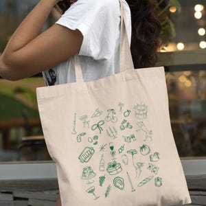 May include: A beige canvas tote bag with green illustrations of various objects, including a wine bottle, a glass of wine, a baguette, a bow, and a tomato.