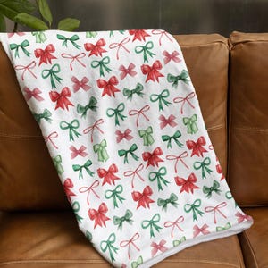 May include: A white blanket with a repeating pattern of red and green bows. The bows are in a watercolor style, with varying shades of red and green. The blanket is folded over a brown leather couch.