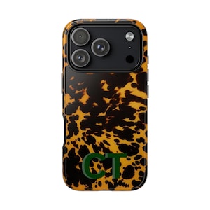 May include: A phone case with a tortoiseshell pattern in shades of brown and yellow. The case has a black camera module with three lenses and a flash. Green initials "CT" are at the bottom of the case.