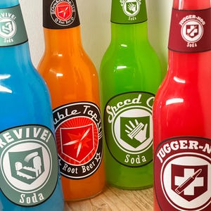 May include: Four brightly coloured soda bottles with distinctive labels. The bottles are blue, orange, green, and red, each featuring a different logo and the words "Revive!", "Double Tap Root Beer", "Speed Cola", and "Juggernog". The bottles are on a wooden surface.