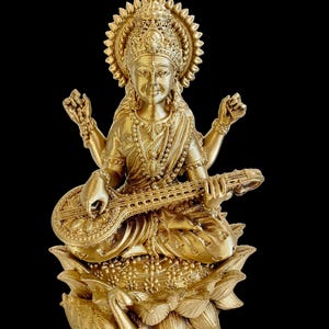 May include: A gold-colored statue of a Hindu goddess playing a stringed instrument. She is seated on a lotus flower with a swan at the base.