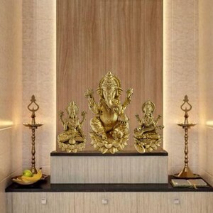 May include: A gold-toned Hindu deity statue set, featuring Ganesha, Lakshmi, and Saraswati, each seated on a lotus base. The statues are arranged on a wooden shelf with two brass oil lamps.