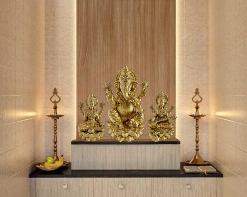 May include: A gold-toned Hindu deity statue set, featuring Ganesha, Lakshmi, and Saraswati, each seated on a lotus base. The statues are arranged on a wooden shelf with two brass oil lamps.
