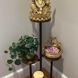 May include: A three-tiered plant stand with a black metal frame and wooden shelves. The top shelf features a gold statue of Ganesha, the second shelf has a potted plant, and the bottom shelf has a decorative bowl with a purple flower.