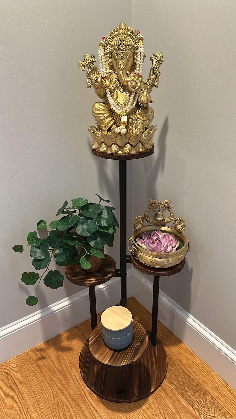 May include: A three-tiered plant stand with a black metal frame and wooden shelves. The top shelf features a gold statue of Ganesha, the second shelf has a potted plant, and the bottom shelf has a decorative bowl with a purple flower.