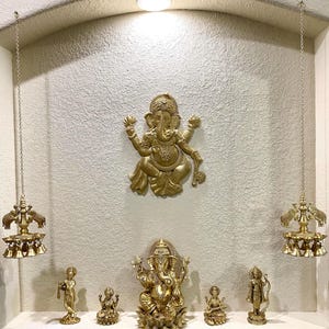 May include: A gold-toned Hindu shrine with a large Ganesha statue in the center. Smaller statues of deities are arranged below, including a Ganesha, a woman playing a musical instrument, and two other figures. Two hanging bells with elephant heads are on either side of the shrine.