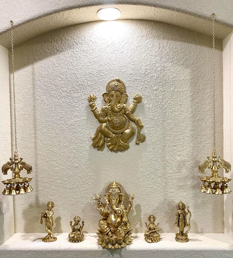 May include: A gold-toned Hindu shrine with a large Ganesha statue in the center. Smaller statues of deities are arranged below, including a Ganesha, a woman playing a musical instrument, and two other figures. Two hanging bells with elephant heads are on either side of the shrine.