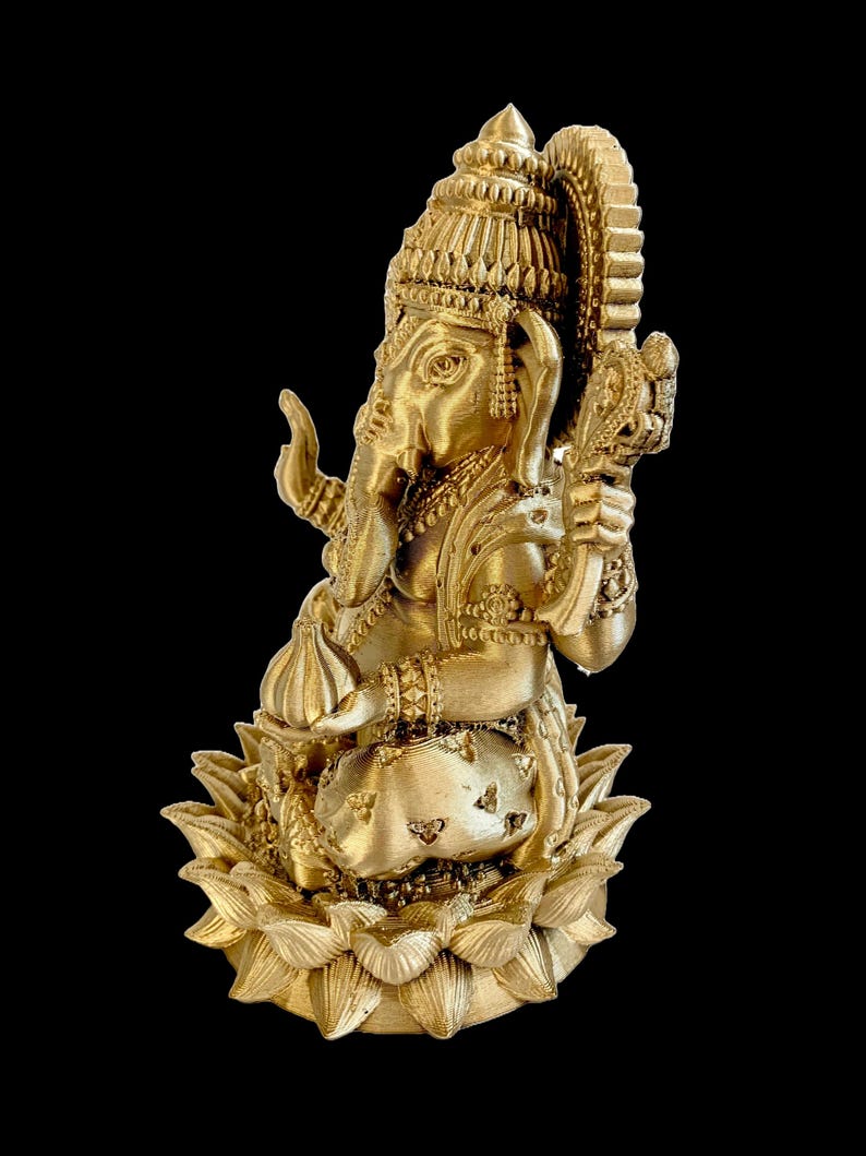 May include: A gold-colored statue of Ganesha, the Hindu god of wisdom and prosperity, sitting on a lotus flower base. The statue is intricately detailed with ornate jewelry and a crown.