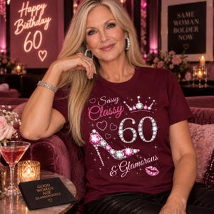 Può includere: Una t-shirt a maniche corte color bordeaux con una stampa grafica che riporta il testo Sassy Classy & Glamorous, il numero 60 tempestato di diamanti, una scarpa con tacco alto decorata con gioielli, una corona e un paio di labbra scintillanti.