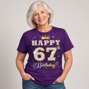 May include: A purple short-sleeved t-shirt featuring a graphic print with a gold crown icon above the text HAPPY 67th Birthday. The numbers 67 are rendered in a silver glitter texture with gold outlines, surrounded by small white starburst accents.