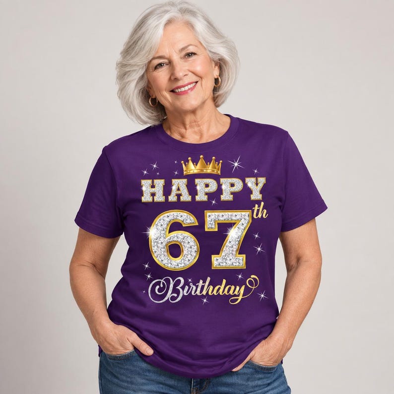 May include: A purple short-sleeved t-shirt featuring a graphic print with a gold crown icon above the text HAPPY 67th Birthday. The numbers 67 are rendered in a silver glitter texture with gold outlines, surrounded by small white starburst accents.