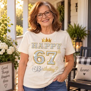 May include: A cream-colored short-sleeve t-shirt featuring a gold crown graphic above the text "HAPPY 67th Birthday" in a silver glitter-style font with gold outlines, accented by small star shapes on the front of the garment.