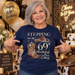 May include: A navy blue t-shirt featuring a graphic print of a leopard print cowboy hat and boot, the number 69 in a jeweled style, and the text "STEPPING Into My 69th Birthday With God's Grace & Mercy" in gold and white lettering.