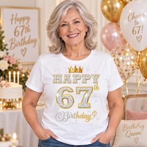 May include: A white short-sleeved t-shirt featuring a gold crown graphic above the text Happy 67th Birthday. The lettering is rendered in a silver glitter texture with gold outlines, accented by small star shapes and a cursive script for the word Birthday.