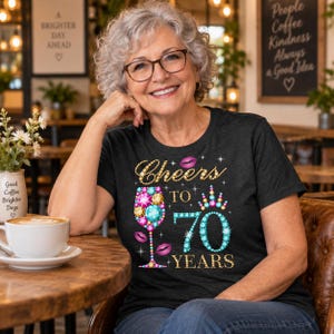 May include: A dark gray short-sleeve t-shirt featuring a graphic print of a wine glass filled with colorful gemstones, a crown, and pink lips. The text reads Cheers TO 70 YEARS in gold and blue lettering with sparkling accents throughout the design.