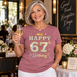 May include: A mauve short-sleeved t-shirt featuring a graphic print with a gold crown and the text "HAPPY 67th Birthday" in a sparkling silver and gold diamond-like pattern, accented with small white starbursts across the front design.