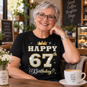 May include: A black short-sleeved t-shirt featuring a graphic design with a gold crown above the text HAPPY 67th Birthday. The numbers 67 are rendered in a silver glitter texture with gold outlines, surrounded by small white star accents on the fabric.