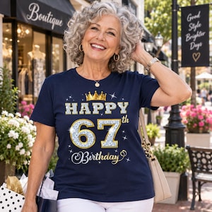 May include: A navy blue short-sleeved t-shirt featuring a graphic print with a gold crown above the text "HAPPY" and the number "67th" in a silver glitter texture, followed by the word "Birthday" in a white script font with star accents.