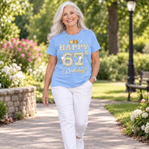 May include: A light blue short-sleeve t-shirt featuring a gold crown graphic above the text "HAPPY 67th Birthday" in a glittery, diamond-patterned font. The design includes small white star accents scattered around the central text graphic.