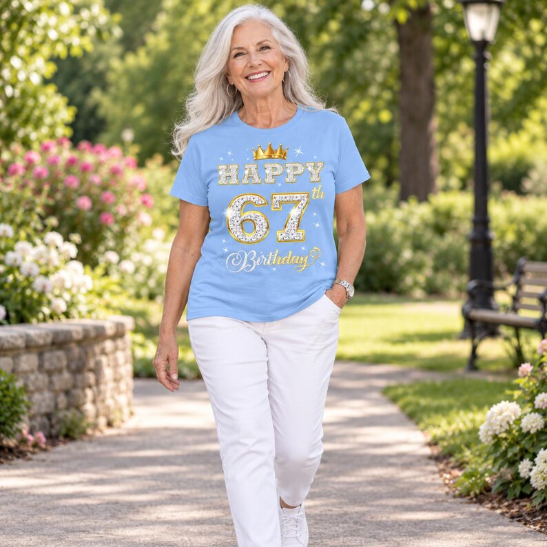 May include: A light blue short-sleeve t-shirt featuring a gold crown graphic above the text "HAPPY 67th Birthday" in a glittery, diamond-patterned font. The design includes small white star accents scattered around the central text graphic.