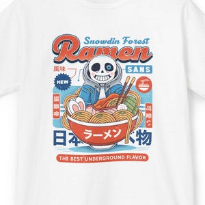 May include: White t-shirt featuring a cartoon illustration of a skeleton character in a bowl of ramen. The design includes the text "Snowdin Forest Ramen" in red and blue, with Japanese characters and the phrase "The Best Underground Flavor."