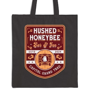 May include: Black tote bag featuring a rectangular emblem with the text "Hushed Honeybee Bar & Inn" in a vintage style. The emblem includes a barrel illustration and the words "Capital Grand Trad" and "Est. 2024".