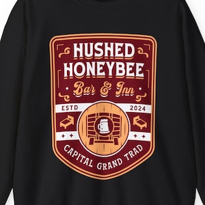 May include: Black sweatshirt with a burgundy, cream, and gold graphic. The graphic reads "Hushed Honeybee Bar & Inn" with a barrel and mug illustration. The text "Capital Grand Trad" is at the bottom, and "Est. 2024" is at the top.