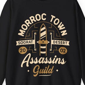 May include: Black sweatshirt with a tan graphic that reads "Morroc Town Desert Assassins Guild." The design includes a stylized dagger and the numbers "20" and "02." The sweatshirt is a crew neck style.