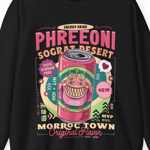 May include: Black sweatshirt featuring a colorful graphic of an energy drink can with a cartoon monster. The can is labeled "PHREEONI SOGRAT DESERT" and "MORROC TOWN Original Flavor". Additional text includes "100% Sugar Free" and Japanese characters.