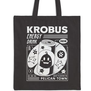 May include: Black tote bag featuring a white graphic design for "Krobus Energy Drink." The design includes a cartoon can, ghost-like characters, and the text "Best Flavor in Pelican Town."