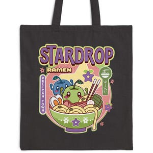 May include: Black tote bag with a colourful graphic of a ramen bowl, the text "STARDROP RAMEN" in purple and yellow, and the words "PELICAN TOWN" printed vertically. The bag has two handles and a cartoon design.