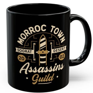 May include: Black ceramic coffee mug with the words "Morroc Town Desert" in a circular design. The mug features a stylized emblem with the text "Assassins Guild" in a gold color. The mug has a black handle and a white background.