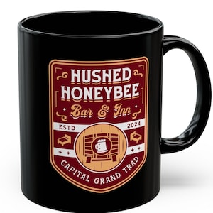 May include: Black ceramic mug with a red and gold logo that reads "Hushed Honeybee Bar & Inn" with the text "ESTD 2024 CAPITAL GRAND TRAD" around the logo.