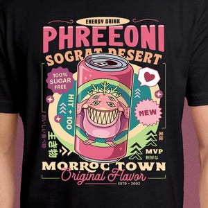May include: Black t-shirt featuring a cartoon graphic of a pink energy drink can with a monster illustration. The can reads "PHREEONI SOGRAT DESERT" and "MORROC TOWN Original Flavor". Additional text includes "100% Sugar Free" and "ESTD-2002".