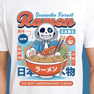 May include: White t-shirt with a cartoon ramen bowl design. The illustration includes a character in a blue hoodie. Text on the shirt reads "Snowdin Forest Ramen Sans" and "The Best Underground Flavor."