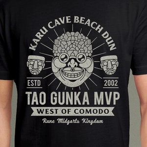 May include: Black t-shirt with a white graphic design featuring a stylized face with a crown of spikes and the text "KARU CAVE BEACH DUN", "ESTD 2002", "TAO GUNKA MVP", "WEST OF COMODO", and "Rune Midgarts Kingdom".