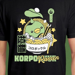 Puede incluir: Una camiseta negra con un diseño gráfico de un personaje de dibujos animados verde en un cuenco de fideos ramen. El texto "KORPORamen" está impreso en la camiseta. El personaje lleva un sombrero verde y tiene un pico amarillo. El cuenco de fideos ramen tiene un borde amarillo y está lleno de fideos, una verdura verde y una gamba roja. El texto "コロポックル" está impreso en el cuenco.