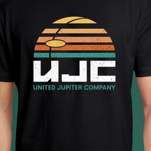 Op de afbeelding: Een zwart T-shirt met korte mouwen en een afbeelding van een zonsondergang in horizontale strepen van geel, oranje en groenblauw. Onder de zonsondergang staan de witte letters UJC, gevolgd door de tekst UNITED JUPITER COMPANY in groenblauw.