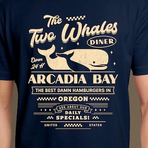 Puede incluir: Camiseta azul marino con un diseño de restaurante de estilo vintage. El diseño presenta el texto "The Two Whales Diner" con ilustraciones de ballenas y las palabras "Arcadia Bay, Oregon". También incluye "Open 24 H" y "Ask about our daily specials!"