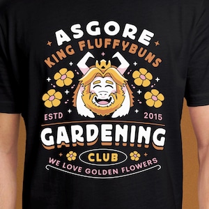 May include: Black t-shirt with a graphic that reads "ASGORE KING FLUFFYBUNS GARDENING CLUB" and "WE LOVE GOLDEN FLOWERS." The design includes a cartoon character with a crown and flowers. The shirt is labelled "ESTD 2015."