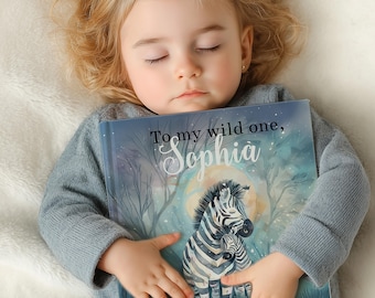 May include: A child is asleep, holding a personalized book. The book cover features a zebra illustration under a full moon, with the text "To my wild one, Sophia" in elegant script. The child is wearing a gray sweater.