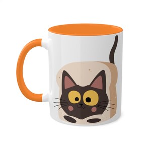 Puede incluir: Taza de cerámica blanca con borde naranja. La taza presenta un gato negro con ojos amarillos asomándose de una rebanada de pan.