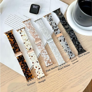 Könnte beinhalten: Eine Auswahl an Apple Watch-Armbändern in verschiedenen Farben und Mustern, darunter Schildpatt, Nougat, rosa Marmor, Perlweiß, Karamell-Schildpatt und schwarzer Marmor. Die Armbänder liegen auf einer Holzoberfläche.